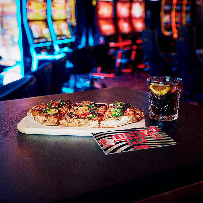 Lucky Me Gutschein, Pinsa, Softdrink auf einem Tisch in der Casino Bar vor bunten Spielautomaten