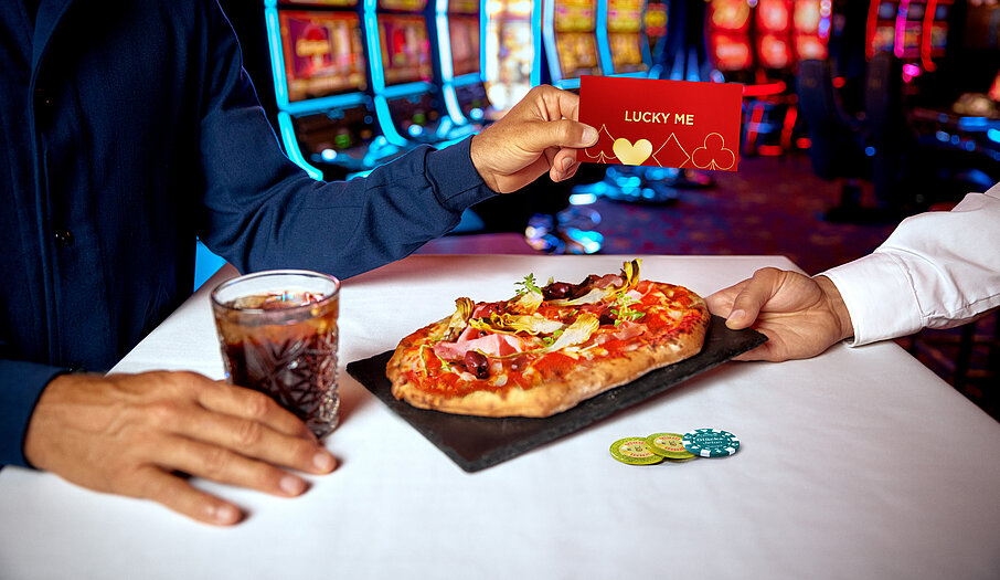 Lucky Me Gutschein, Pinsa, Softdrink auf einem Tisch in der Casino Bar vor bunten Spielautomaten