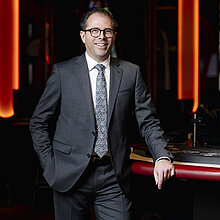 Marketing Expert im Casino Graz, Stefan Bachernegg, trägt einen grauen Anzug mit gemusterter Krawatte und steht an einem Pokertisch, lächelnd in die Kamera.