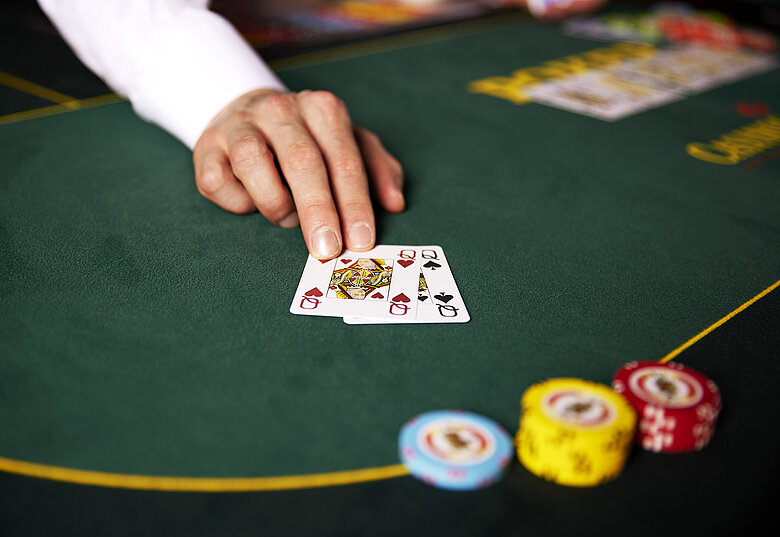 Hand von Croupier schiebt zwei Damen Poker Karten über den grünen Tisch, daneben bunte gestapelte Jetons und verschiedene Karten