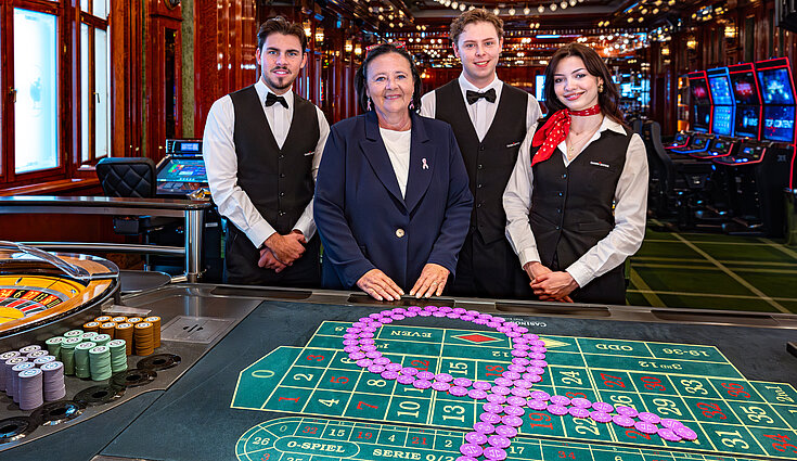 Casino-Mitarbeiter mit Casino-Managerin vor Roulettetisch mit pinken Chips in Form eines Bandes
