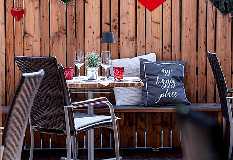 Gemütliche Terrasse mit Holzwand, roter Herzdeko und gedecktem Tisch. Rattanstühle und Kissen schaffen eine einladende Atmosphäre.