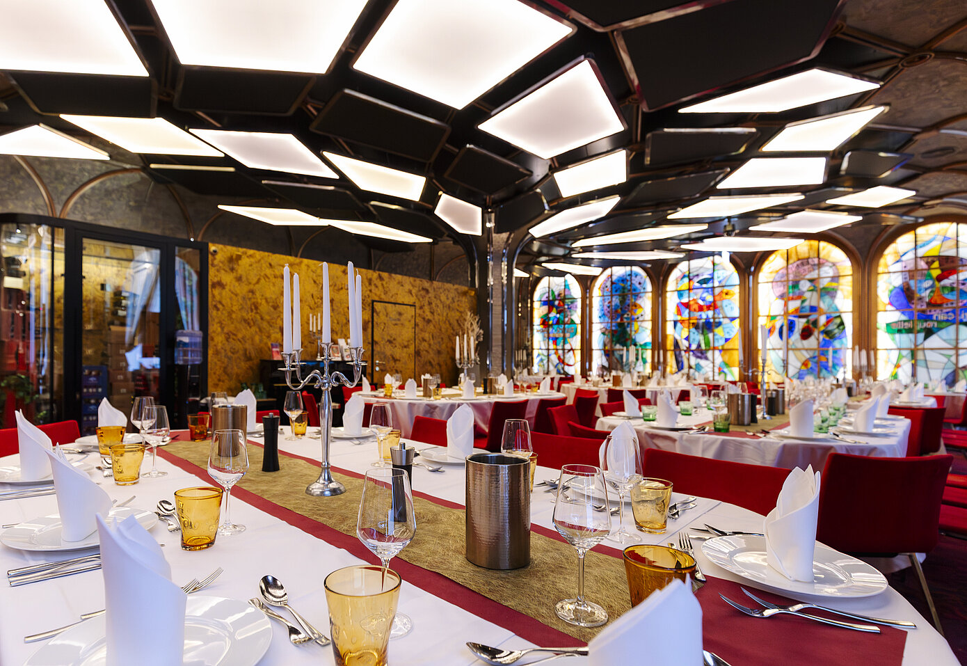 Eleganter Restaurantinnenraum mit gedeckten Tischen. Die Tische sind mit weißen Tischdecken, roten Tischläufern, gelben Gläsern und silbernen Kerzenständer eingedeckt. Im Hintergrund sind bunte Glasfenster und goldene Vertäfelungen zu sehen.