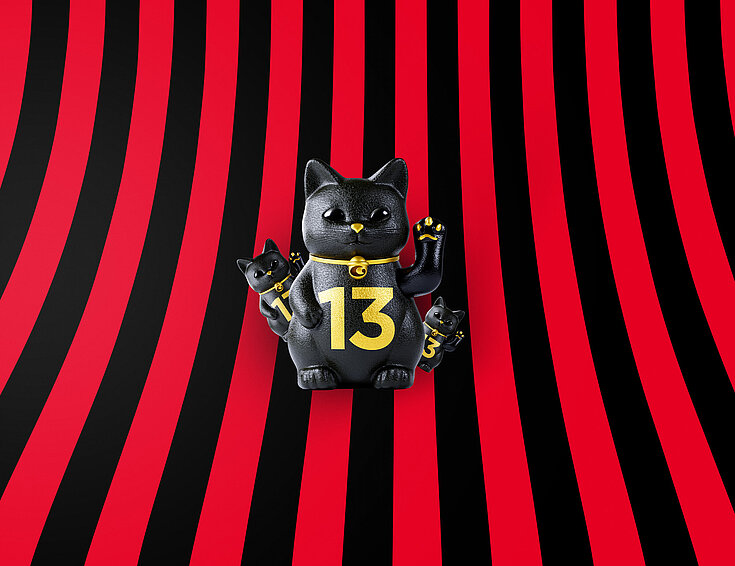 Eine große und zwei kleine schwarze Glückskatzen mit goldener 13 auf rot-schwarz-gestreiftem Hintergrund
