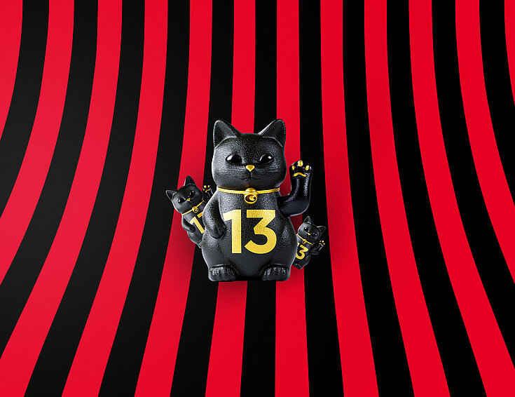 Eine große und zwei kleine schwarze Glückskatzen mit goldener 13 auf rot-schwarz-gestreiftem Hintergrund