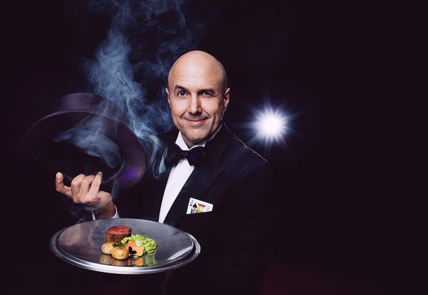 Magier Jack Black in einem Smoking, präsentiert ein dampfendes Gericht auf einem Tablett, während er mit einer Hand einen Zaubertrick ausführt.