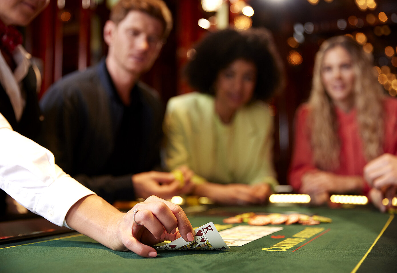 Hand von Croupier deckt Karte König auf einem Pokertisch auf, dahinter drei Personen, die gespannt die Karte betrachten
