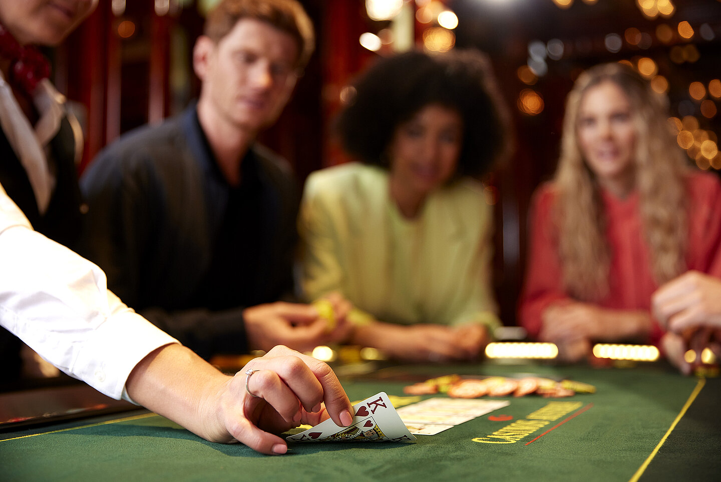 Hand von Croupier deckt Karte König auf einem Pokertisch auf, dahinter drei Personen, die gespannt die Karte betrachten