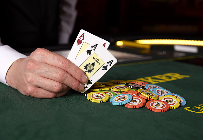 Hand von Croupier liegt auf Pokertisch, hält zwei Asse hoch, nebenbei bunte Jetons