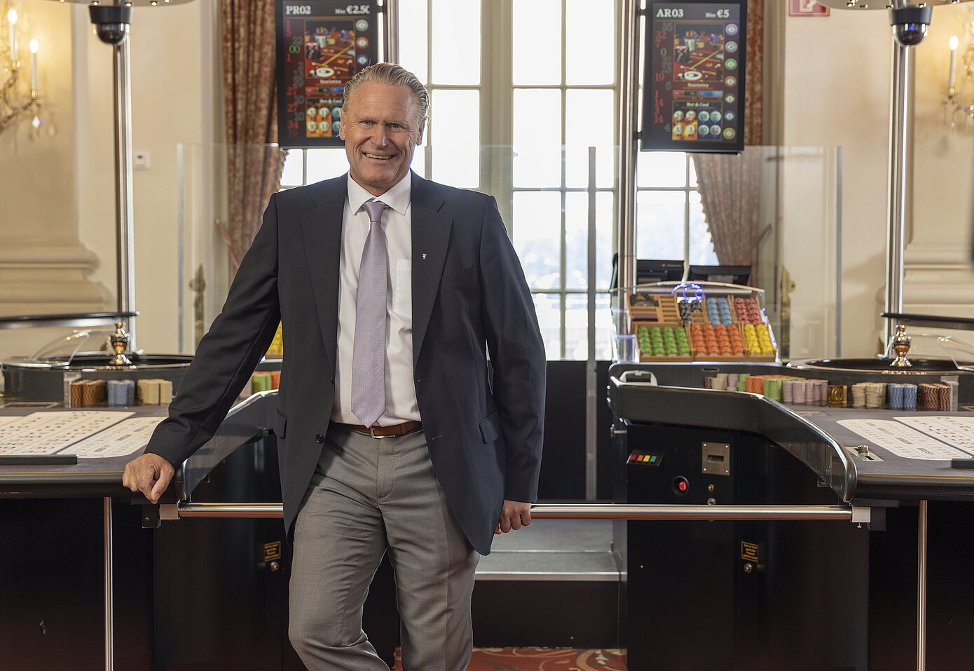 Mann mittleren Alters, helle Haare zurückgekämmt, mit weißem Hemd, blauer Krawatte, grauer Anzugshose und schwarzem Sacko stehend zwischen zwei Spieltischen im Casino Zell am See