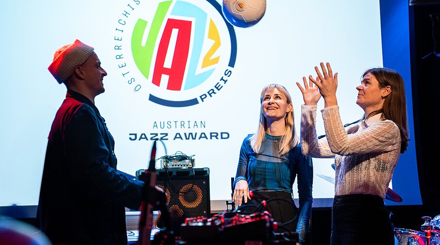 Drei Menschen stehen auf einer Bühne vor Musikinstrumenten