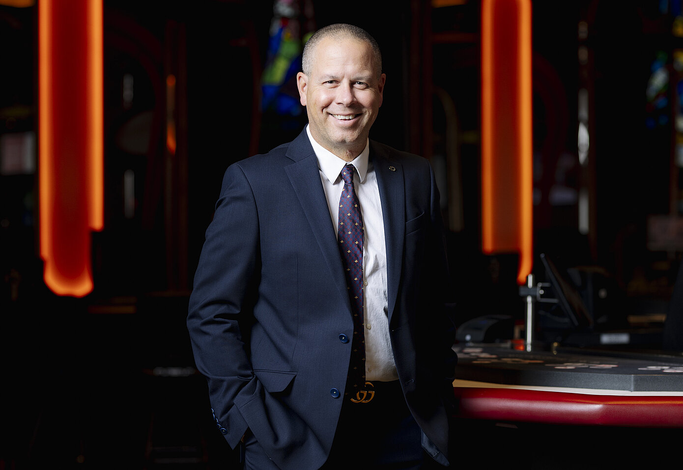 Casino Direktor im Casino Graz, Manuel Haderer, trägt einen blauen Anzug mit lila Krawatte und steht an einem Pokertisch, lächelnd in die Kamera.