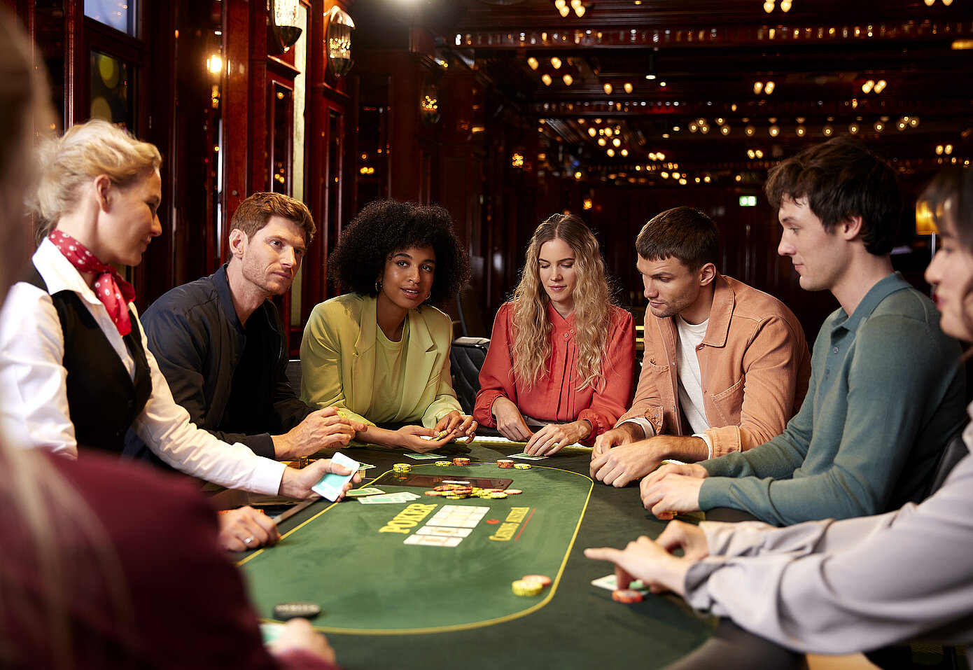Gruppe verschiedenster Menschen sitzend um Pokertisch, gespannte Blicke auf Spiel, Croupier hat Karten in der Hand, Jetons am Tisch