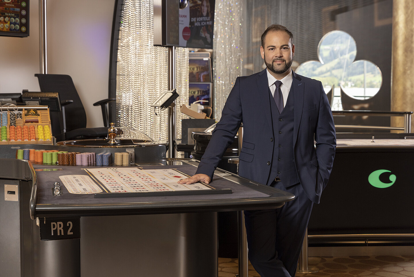 Mann mit schwarzen Haaren und Vollbart, in dunkelblauem Anzug stehend zwischen zwei Spieltischen im Casino Zell am See
