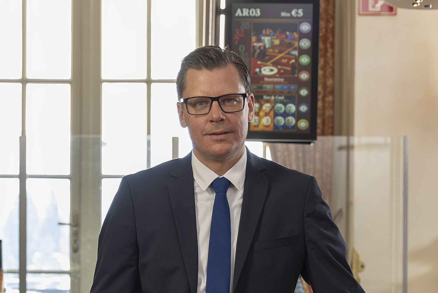 Mann Mitte 40 mit dunklen Haaren und schwarzer Brille in einem dunkelblauen Anzug mit blauer Krawatte